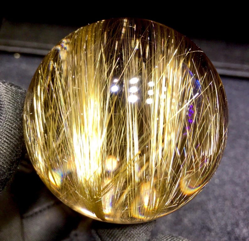 Puede incluir: Una bola de cristal transparente con inclusiones doradas en forma de aguja. Las inclusiones crean un efecto visual llamativo, que se asemeja a hebras de oro. El cristal refleja la luz, creando una exhibici&oacute;n brillante e iridiscente. Un art&iacute;culo de decoraci&oacute;n &uacute;nico.