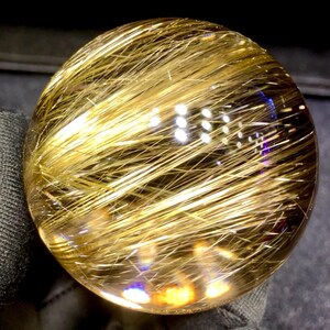 Puede incluir: Una bola de cristal transparente y esf&eacute;rica con inclusiones doradas en forma de cabello. Las inclusiones est&aacute;n densamente empaquetadas e irradian por toda la esfera, creando un efecto brillante. El cristal es liso y pulido, reflejando la luz.