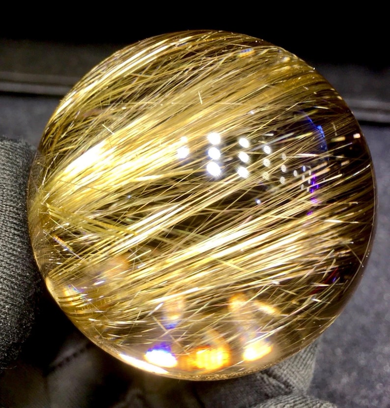 Puede incluir: Una bola de cristal transparente y esf&eacute;rica con inclusiones doradas en forma de cabello. Las inclusiones est&aacute;n densamente empaquetadas e irradian por toda la esfera, creando un efecto brillante. El cristal es liso y pulido, reflejando la luz.