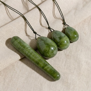 May include: A collection of green jade products, including three egg-shaped pendants and one cylindrical wand. Each piece is attached to a dark green cord. The items are displayed on a beige fabric surface.