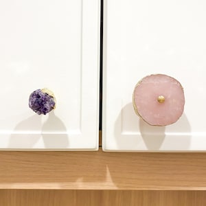 Puede incluir: Dos pomos de armario con un borde dorado. Un pomo está hecho de un cristal de amatista púrpura y el otro de un ágata rosa.