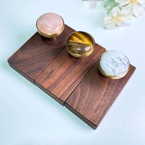 Puede incluir: Tres ganchos redondos decorativos con incrustaciones de piedra diferentes. Los ganchos están montados sobre una base de madera. Las piedras son rosas, marrones y blancas.