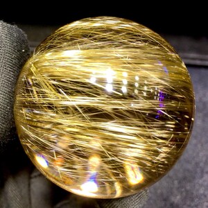 Puede incluir: Una bola de cristal transparente y esf&eacute;rica con inclusiones doradas en forma de aguja. Las inclusiones crean un efecto brillante, reflejando la luz y dando a la esfera una apariencia radiante. El cristal est&aacute; pulido y liso, con un ligero tinte amarillo.