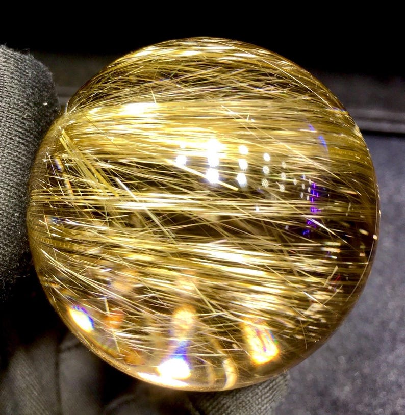 Puede incluir: Una bola de cristal transparente y esf&eacute;rica con inclusiones doradas en forma de aguja. Las inclusiones crean un efecto brillante, reflejando la luz y dando a la esfera una apariencia radiante. El cristal est&aacute; pulido y liso, con un ligero tinte amarillo.
