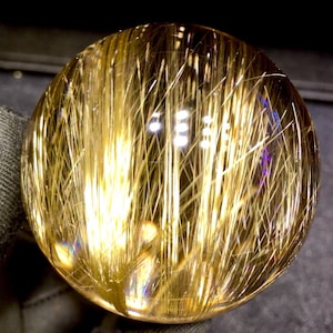 Puede incluir: Una bola de cristal transparente y esf&eacute;rica con inclusiones doradas en forma de aguja. Las inclusiones est&aacute;n densamente empaquetadas, creando un efecto visual llamativo. La bola est&aacute; pulida y refleja la luz, mostrando su estructura interna y variaciones de color.