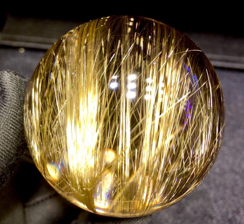 Puede incluir: Una bola de cristal transparente y esf&eacute;rica con inclusiones doradas en forma de aguja. Las inclusiones est&aacute;n densamente empaquetadas, creando un efecto visual llamativo. La bola est&aacute; pulida y refleja la luz, mostrando su estructura interna y variaciones de color.
