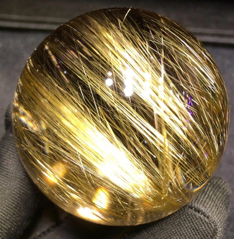 Puede incluir: Una esfera de cristal transparente con inclusiones doradas en forma de aguja. La esfera es clara, lo que permite que la luz la atraviese y resalte los hilos dorados. El cristal parece pulido y liso, reflejando la luz.