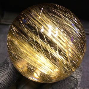 Puede incluir: Una bola de cristal esf&eacute;rica y transparente con inclusiones doradas en forma de aguja. Las inclusiones est&aacute;n densamente empaquetadas y crean un efecto brillante. La bola se sostiene sobre un fondo oscuro, resaltando su claridad y estructura interna.