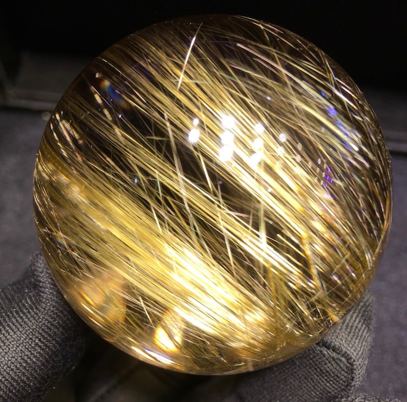 Puede incluir: Una bola de cristal esf&eacute;rica y transparente con inclusiones doradas en forma de aguja. Las inclusiones est&aacute;n densamente empaquetadas y crean un efecto brillante. La bola se sostiene sobre un fondo oscuro, resaltando su claridad y estructura interna.