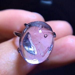Könnte beinhalten: Ein Ring mit einem großen, ovalen, durchscheinenden hellvioletten Edelstein mit sichtbaren Einschlüssen und Blasen im Inneren. Der Stein ist in einer einfachen Metallfassung mit vier Krappen befestigt und hat eine glatte, polierte Oberfläche.