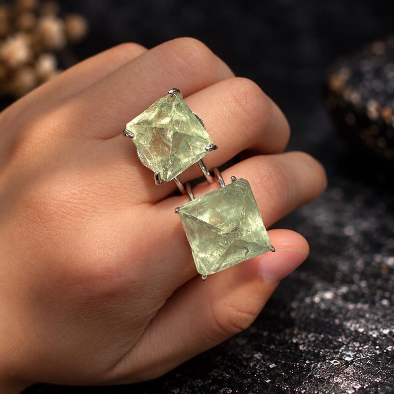 K&ouml;nnte beinhalten: Ein Doppelring mit zwei gro&szlig;en, quadratisch geschliffenen, blassgr&uuml;nen, durchscheinenden Edelsteinen, die in silberfarbenen Metallfassungen sitzen. Die Steine haben eine facettierte, pyramidenartige Struktur mit sichtbaren inneren Texturen.