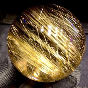Puede incluir: Una bola de cristal transparente con inclusiones doradas en forma de aguja. Las inclusiones est&aacute;n densamente empaquetadas, creando un efecto brillante. El cristal se sostiene en una tela oscura y texturizada, resaltando su brillo. Este art&iacute;culo decorativo es ideal para coleccionistas.