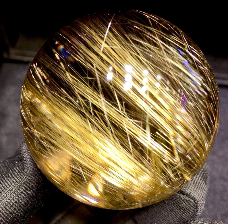 Puede incluir: Una bola de cristal transparente con inclusiones doradas en forma de aguja. Las inclusiones est&aacute;n densamente empaquetadas, creando un efecto brillante. El cristal se sostiene en una tela oscura y texturizada, resaltando su brillo. Este art&iacute;culo decorativo es ideal para coleccionistas.