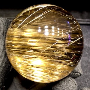 Puede incluir: Una bola de cristal esf&eacute;rica y transparente con inclusiones doradas en forma de aguja. Las inclusiones crean un efecto texturizado llamativo dentro de la esfera transparente. La bola de cristal se sostiene en una tela gris oscura.