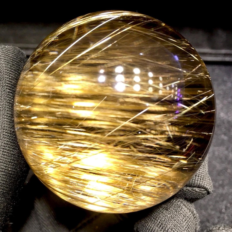 Puede incluir: Una bola de cristal esf&eacute;rica y transparente con inclusiones doradas en forma de aguja. Las inclusiones crean un efecto texturizado llamativo dentro de la esfera transparente. La bola de cristal se sostiene en una tela gris oscura.