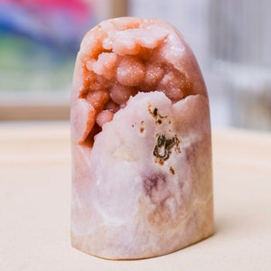 May include: A pink and white geode with a large cluster of crystals inside. The geode is shaped like a rounded rectangle and has a smooth, polished surface.