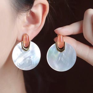 May include: A pair of elegant earrings featuring a large, iridescent white disc with a gold-colored accent and a peach-colored stone. The earrings are designed to be worn on the ear.