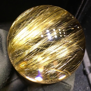 Puede incluir: Una bola de cristal esf&eacute;rica transparente con inclusiones doradas en forma de cabello. Las inclusiones se distribuyen uniformemente por toda la esfera, creando un efecto brillante. La bola de cristal se sostiene sobre un fondo oscuro.