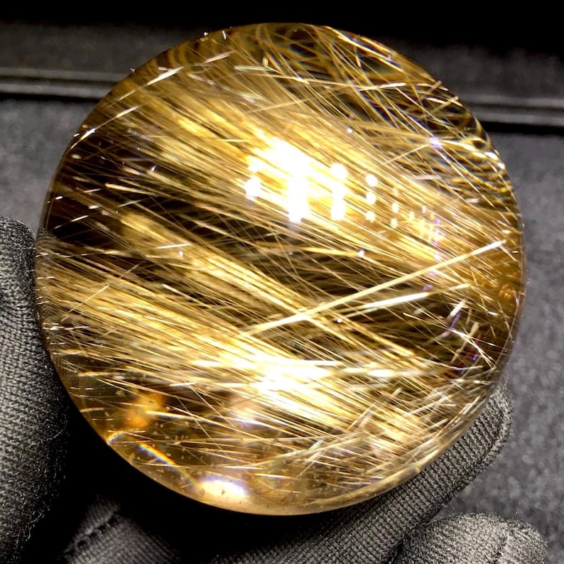 Puede incluir: Una bola de cristal transparente con inclusiones doradas en forma de aguja. Las inclusiones est&aacute;n densamente empaquetadas, creando un efecto brillante. El cristal se sostiene sobre un fondo oscuro, resaltando su claridad y estructura interna.