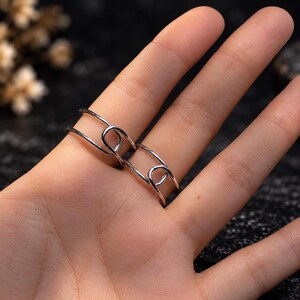 K&ouml;nnte beinhalten: Ein silberfarbener Ring mit einem Doppelband-Design, das die Form einer Sicherheitsnadel nachahmt. Das Metallband umschlie&szlig;t den Finger mit zwei parallelen Linien, die an einem schlaufenf&ouml;rmigen Verschluss auf der Vorderseite verbunden sind.
