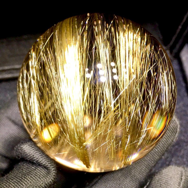 Puede incluir: Una esfera de cristal transparente con inclusiones doradas en forma de aguja. Las inclusiones est&aacute;n densamente empaquetadas, creando un efecto brillante. El cristal se sostiene sobre un fondo oscuro, resaltando su claridad y el color dorado.
