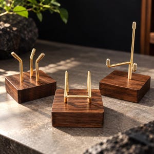 Pode incluir: Três suportes de exibição com bases quadradas de madeira castanho-escura e suportes de metal dourado. Um suporte tem três pontas verticais, um tem um berço em forma de U e o terceiro tem um braço ajustável em forma de L com poste.