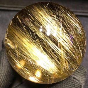 Puede incluir: Una esfera de cristal transparente con inclusiones doradas en forma de aguja. Las inclusiones crean un efecto visual llamativo, que se asemeja a hebras de oro. La esfera est&aacute; pulida y refleja la luz, mostrando su estructura interna y variaciones de color.