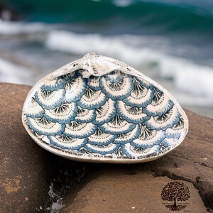 May include: A decorative seashell bowl with a painted interior. The bowl features a white base with a blue and black scalloped pattern. The shell is resting on a rock with the ocean in the background.