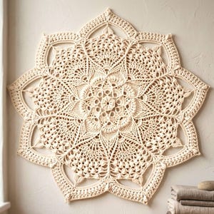 Puede incluir: Un tapiz de pared de ganchillo en forma de estrella, color crema. El intrincado diseño presenta un motivo floral central rodeado de patrones calados detallados. La pieza está montada en una pared de tonos neutros, mostrando su delicada textura y artesanía.
