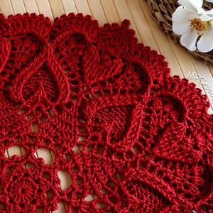 May include: A close-up of a red crocheted doily with a delicate floral pattern. The doily is made with a fine yarn and has a intricate design.