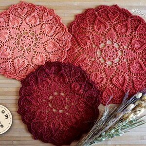 May include: Three crocheted doilies in shades of red and pink. The doilies are circular and feature a delicate, lacy design. The doilies are arranged on a wooden surface with dried flowers.