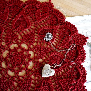 May include: A red crocheted doily with a delicate pattern. A silver chain necklace with a heart-shaped pendant and a jeweled pin are placed on top of the doily.