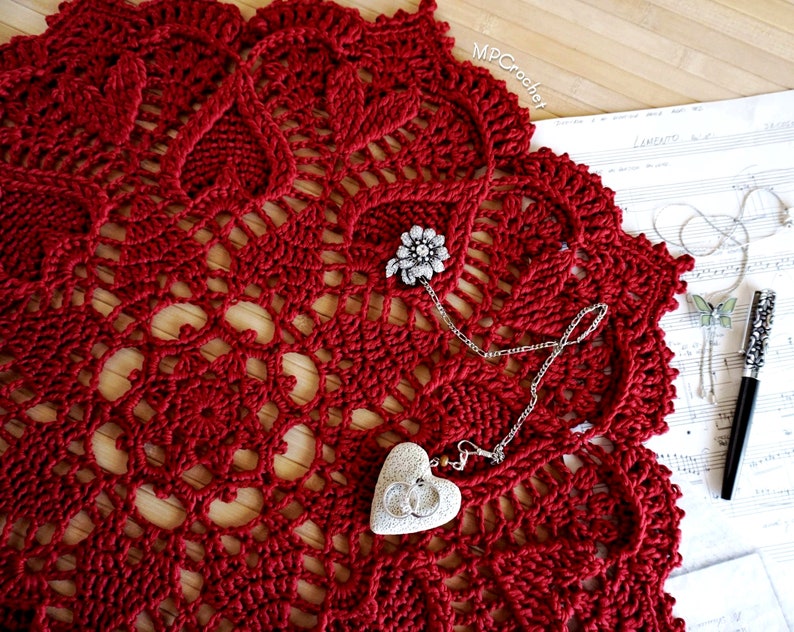 May include: A red crocheted doily with a delicate pattern. A silver chain necklace with a heart-shaped pendant and a jeweled pin are placed on top of the doily.