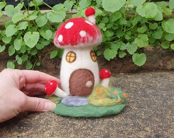 May include: A hand-felted mushroom house with a red and white mushroom cap, a white body with a brown door, and a green base with small mushrooms and flowers.