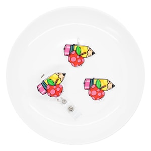May include: Three pencil and apple-themed badge reels on a white surface. The pencil is yellow with a pink eraser and a black tip. The apple is red with pink polka dots and a green leaf. One reel has a clear plastic retractable cord.