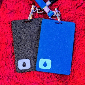 May include: Two rectangular ID badges, one black and one blue, with a glittery texture. Each badge has a small, light blue square with a water droplet icon. A blue lanyard with silver clasps is attached to the badges.