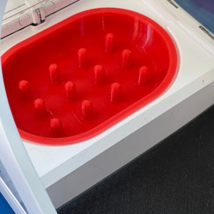 Puede incluir: Una bandeja de plástico blanco con un inserto de silicona rojo. El inserto tiene 12 pequeñas hendiduras redondas.