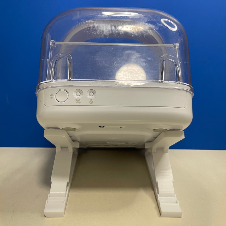 Puede incluir: Dispositivo de pl&aacute;stico blanco con una c&uacute;pula de pl&aacute;stico transparente, dos botones etiquetados como "Izquierda" y "Derecha", y dos patas plegables. El dispositivo est&aacute; dise&ntilde;ado para sostener y mostrar objetos peque&ntilde;os.