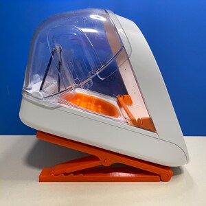 Puede incluir: Un recipiente de comida de pl&aacute;stico blanco y naranja con una tapa transparente. El recipiente tiene un soporte integrado que se pliega para facilitar el acceso.