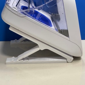 May include: A white plastic device with a clear plastic top and a blue interior. The device has a foldable stand with two legs. The device is designed to hold and dispense water.