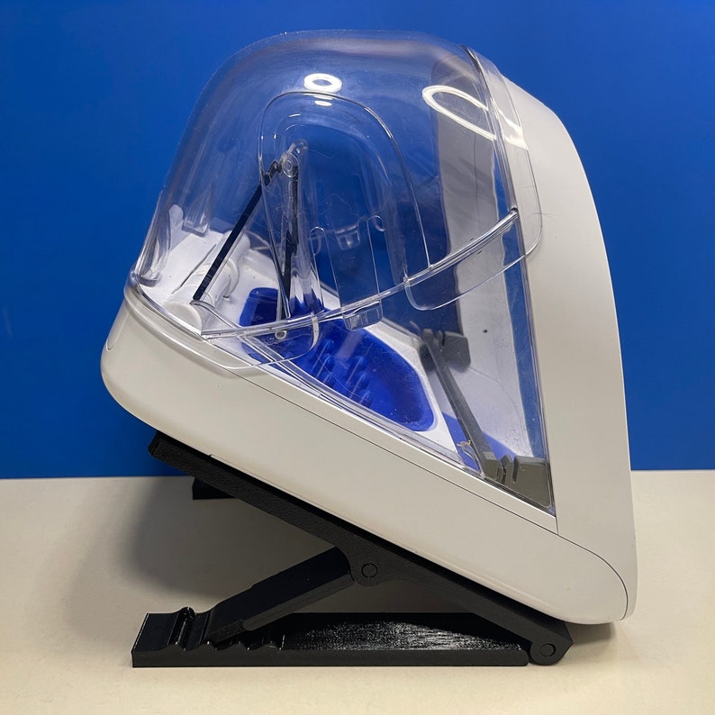 Puede incluir: Un dispositivo de pl&aacute;stico blanco y transparente con un interior azul, sentado en un soporte plegable negro. El dispositivo tiene una tapa transparente y una abertura con bisagras.