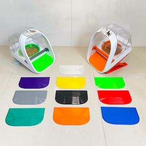 Puede incluir: Dos dispensadores automáticos de comida para mascotas blancos con una base verde y naranja. Los dispensadores están sobre una superficie blanca con 10 alfombras de diferentes colores delante de ellos. Las alfombras son moradas, amarillas, grises, negras, naranjas, rojas, azules, verdes, blancas y verde claro.
