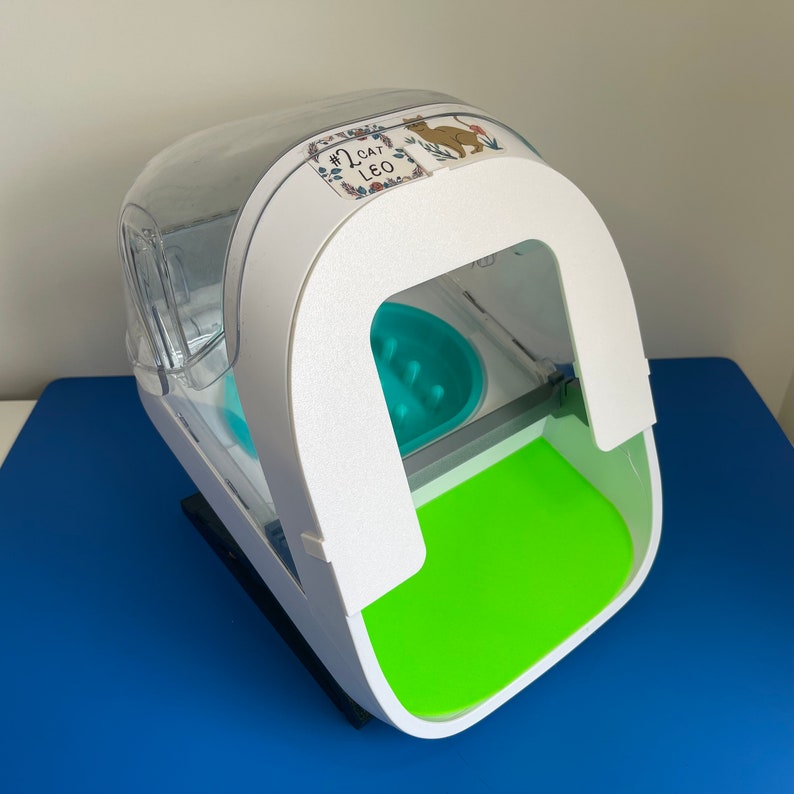 May include: White automatic pet feeder with a clear plastic top and a green food bowl. The feeder has a sticker that says "#1 Cat Leo".