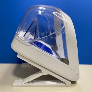 Puede incluir: Un dispositivo de pl&aacute;stico blanco con una c&uacute;pula de pl&aacute;stico transparente en la parte superior. El dispositivo tiene una base plegable y est&aacute; dise&ntilde;ado para sostener un objeto peque&ntilde;o.