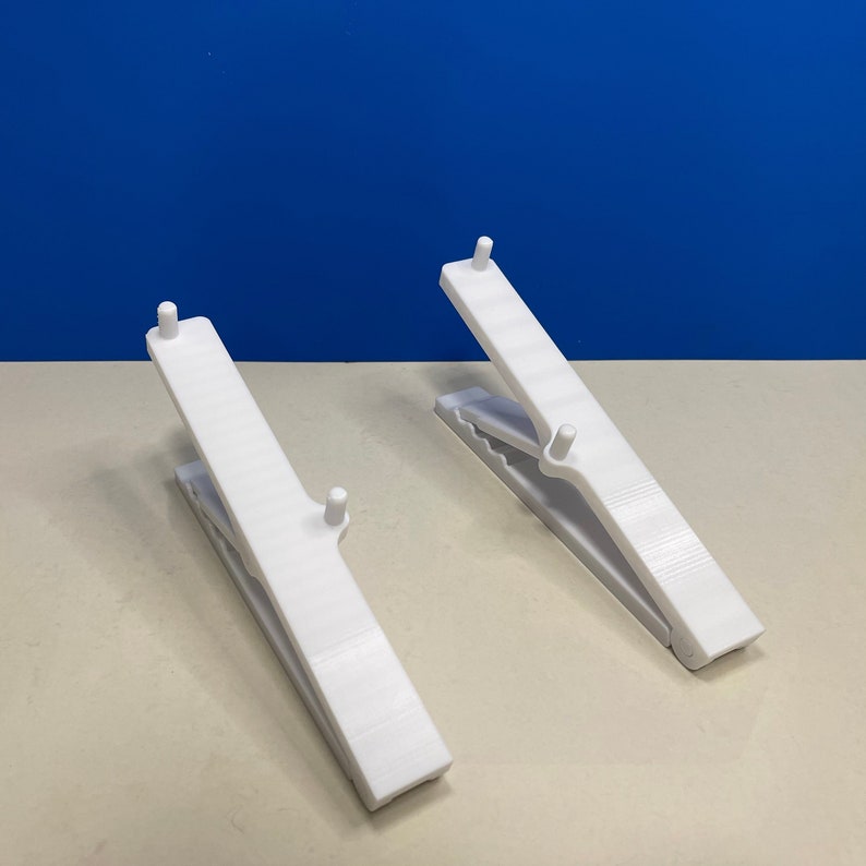 Puede incluir: Dos pinzas de ropa de pl&aacute;stico blanco, cada una con una superficie texturizada y una perilla elevada en la parte superior. Las pinzas de ropa est&aacute;n abiertas y colocadas planas sobre una superficie marr&oacute;n claro.