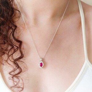 Puede incluir: Un collar de cadena de plata con un colgante que presenta una gran piedra ovalada roja rodeada de piedras pequeñas transparentes.