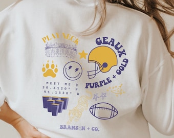 May include: White long-sleeve t-shirt with a purple and gold graphic design. The graphic includes a football helmet, a tiger, a paw print, a smiley face, a star, and the text "Play Neck Geaux Purple + Gold Meet Me @ 30.4120° N 91.1838° W Branson + Co."