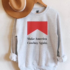 Puede incluir: Sudadera gris con un rect&aacute;ngulo rojo y el texto "Make America Cowboy Again."