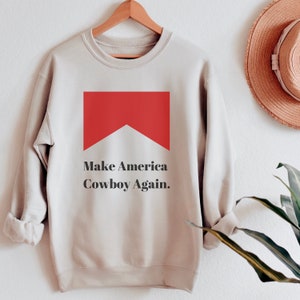 Puede incluir: Una sudadera beige con un gr&aacute;fico rojo y blanco de un paquete de cigarrillos y el texto "Make America Cowboy Again."