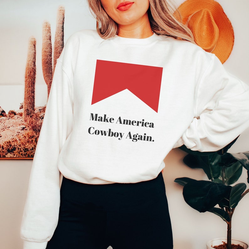 Puede incluir: Sudadera blanca con un gr&aacute;fico rojo de un cigarrillo y el texto "Make America Cowboy Again."